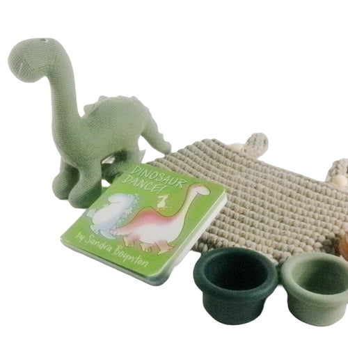 A children's dinosaur-themed toy set on a white background, including a green textured dinosaur figure, a small book titled 'Dinosaur Dance!' by Sandra Boynton, a woven mat, and two small cups in dark green and light green.
