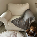 White armchair with a gray blanket, teddy bear, and children's rug in a room.