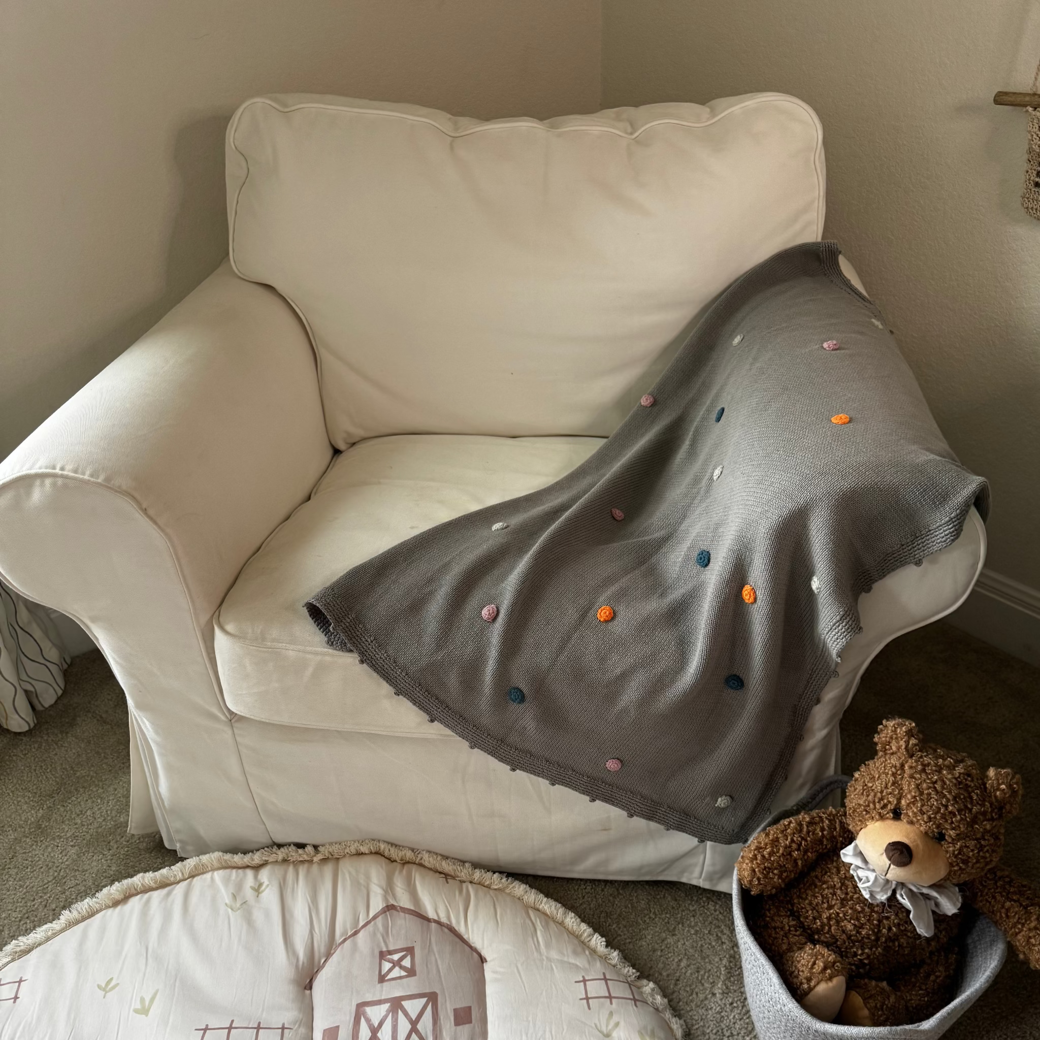 White armchair with a gray blanket, teddy bear, and children's rug in a room.