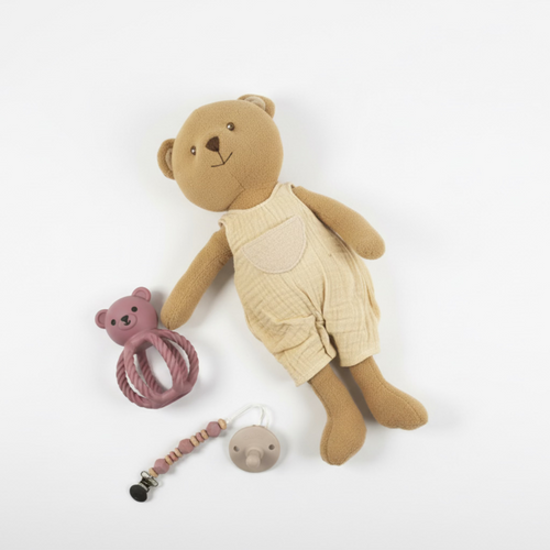Brown teddy bear with beige romper, pink rattle, and pacifier clip on a white background